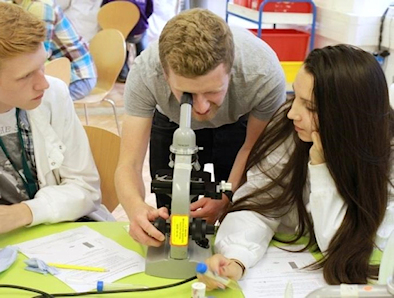 Two students sitting either side of a researcher who looks down a microscope