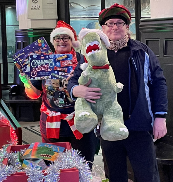 A woman holding 2 selection boxes, standing next to a man holding a large T-rex toy