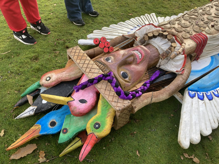 Winged figure with a hat composed of colourful hummingbird heads, carved of wood and laid flat on the ground