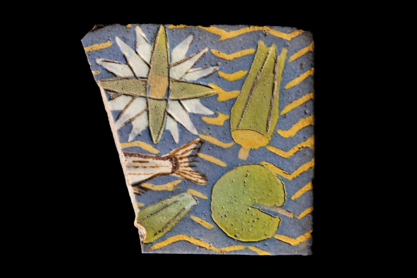 Part of a ceramic tile showing the tail fo a fish, lillypads and other green, white and blue decoration